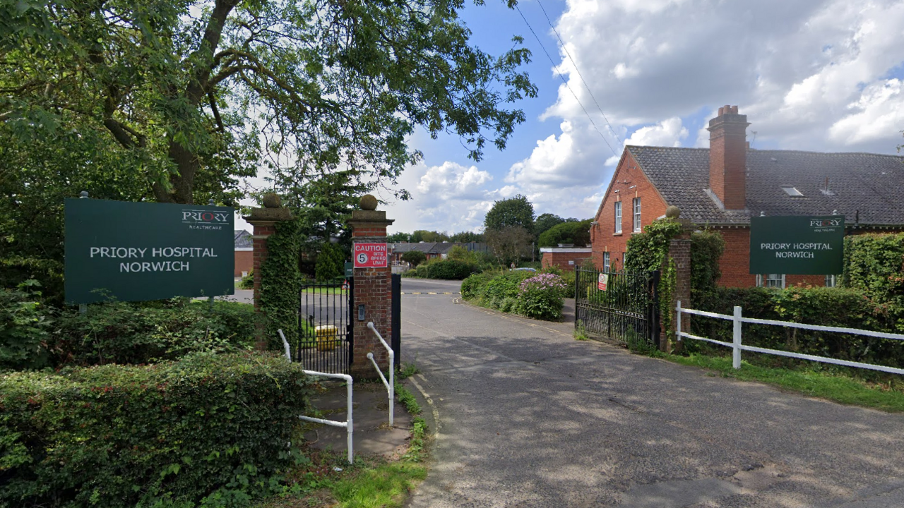 The entrance gate to Priory Hospital Norwich on a clear day with blue skies. A red brick building is on one side of the road with trees on the other.