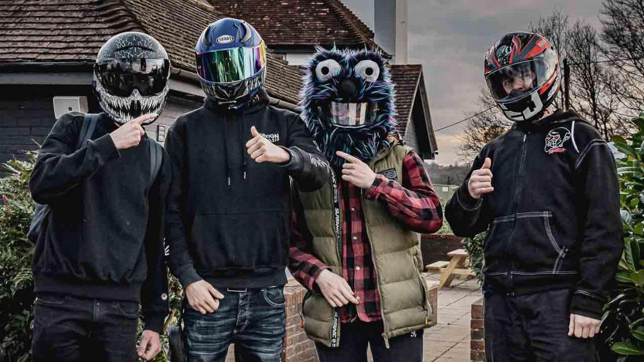 Four youths stand at The Hut wearing motorcycle helmets. One of the helmets is in the style of a furry animal head.