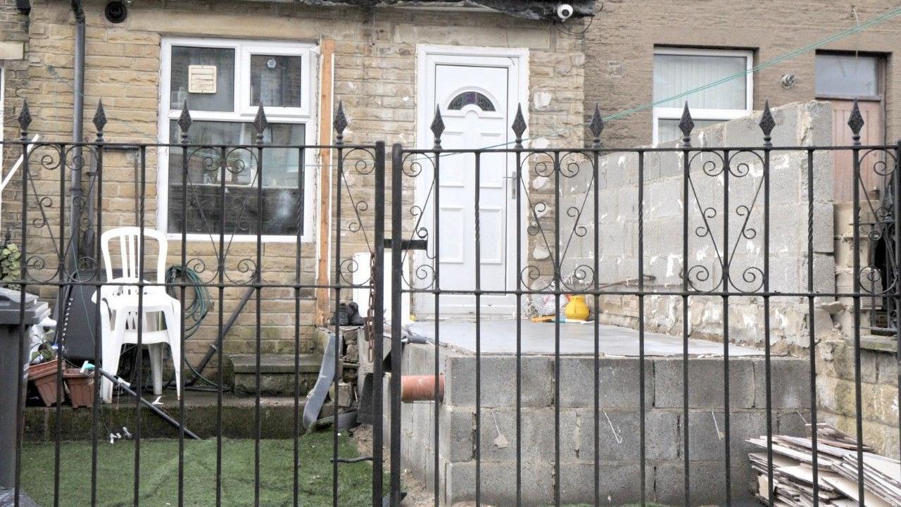 The photograph shows the ongoing work to remove the extension. The door is now visible through a metal fence, with a concrete structure and a side wall remaining.