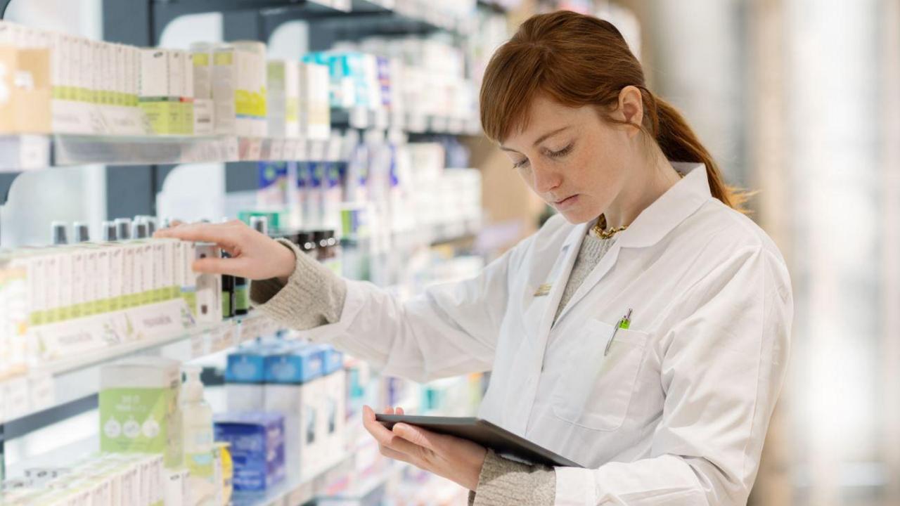 A woman with brown hair in a white pharmacists coat holding a tablet in one hand and reaching for a box of medicine from a shelf with the other