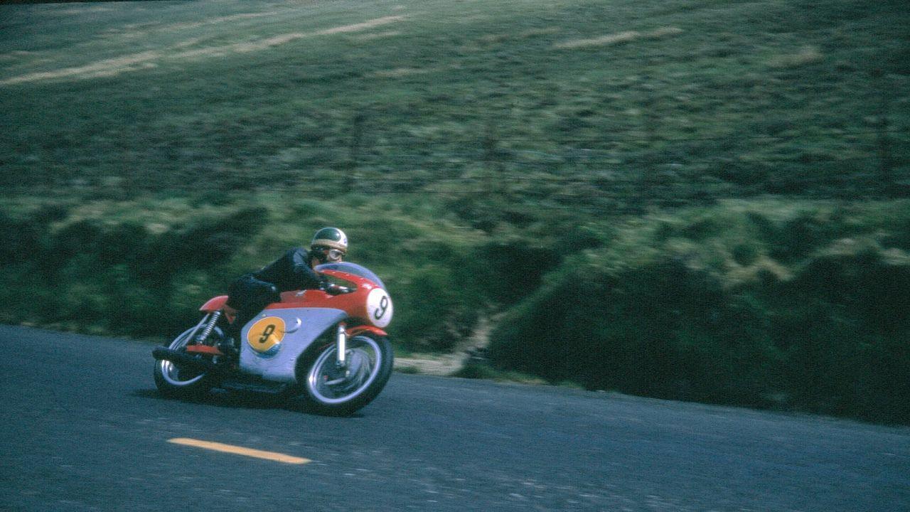 An old-fashioned picture of Giacomo Agostini on a bubble shaped red motorbike, with a number nine as he tilts as he turns a corner on the mountain road.