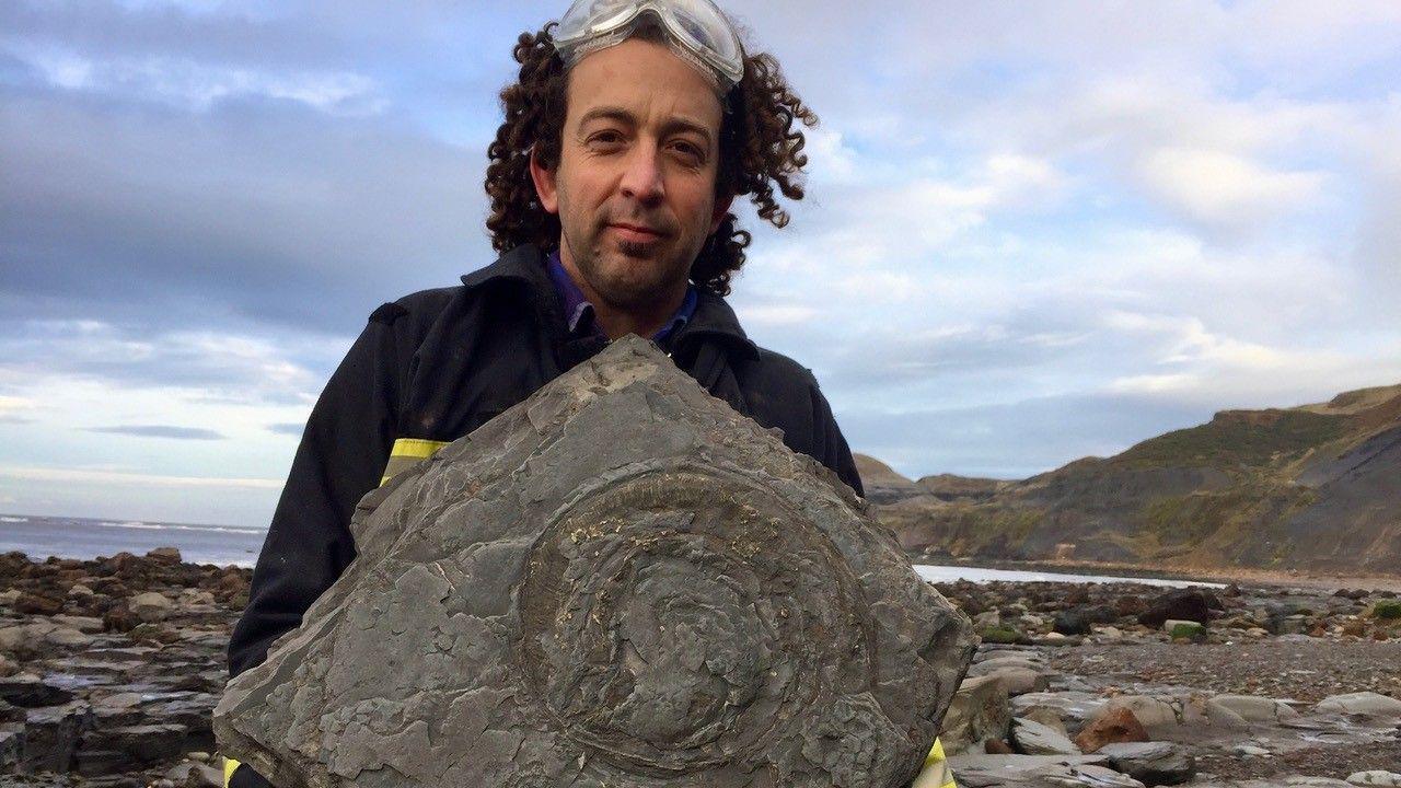 A man with dark curly hair holds up a large rock with the imprint of a fossil in the centre. He has clear plastic glasses on the top of his head, and is dressed in dark workman-style clothes, with bright green gloves. A rocky coastline and the sea can be seen behind him.
