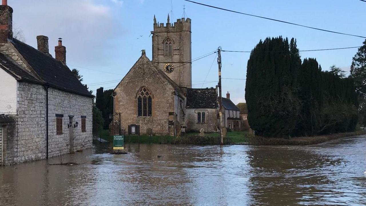 Flood water on a road, and in the background a church. The flood waters appear to reach halfway up a house on the left of the shot.