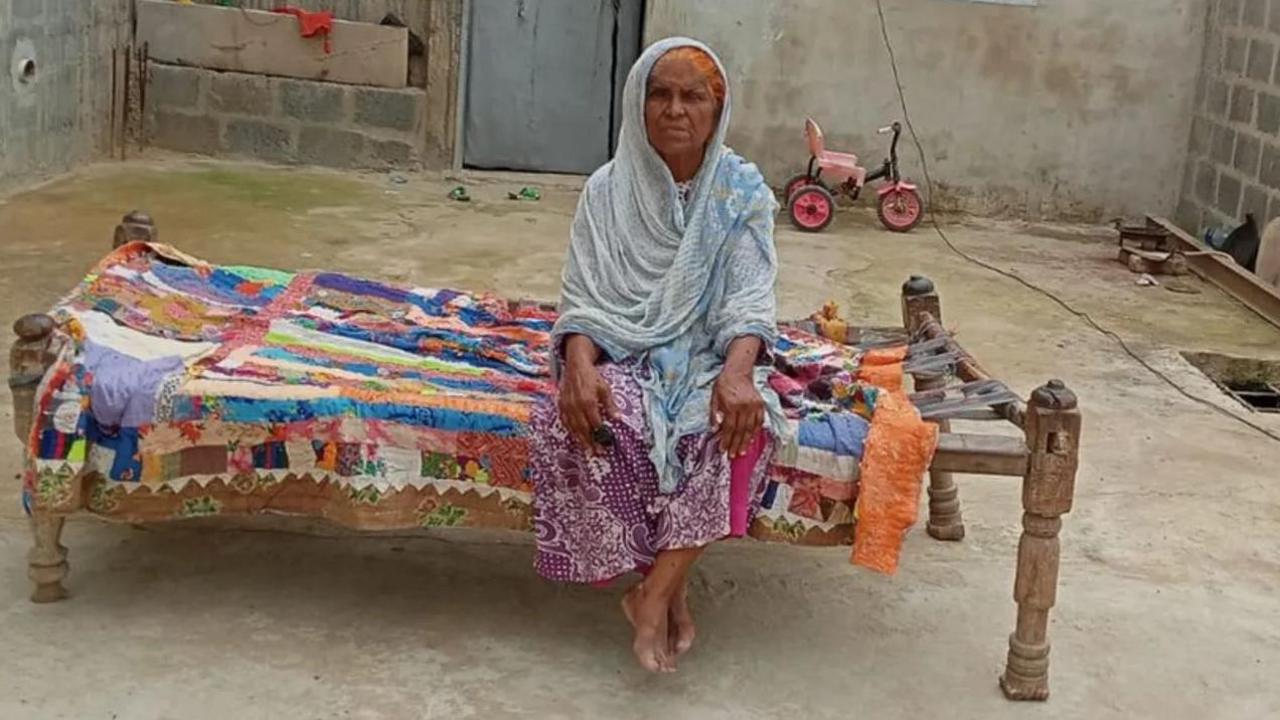 Hamida Banu is sitting on a charpoy in blue-white clothes. She is sitting in a big grey couryard, wrapped in a shawl.