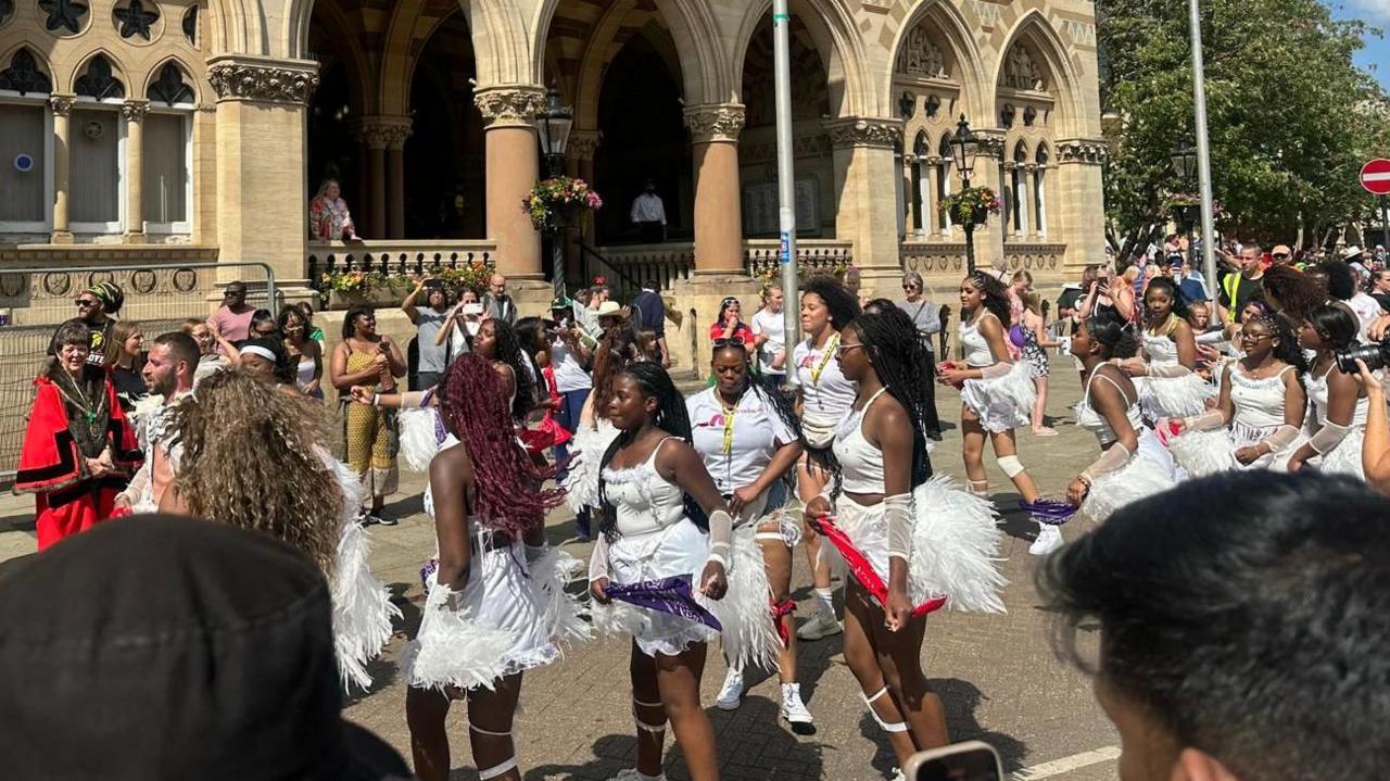 People wearing white tops with fluffy white skirts dancing in front of a grand civic building.