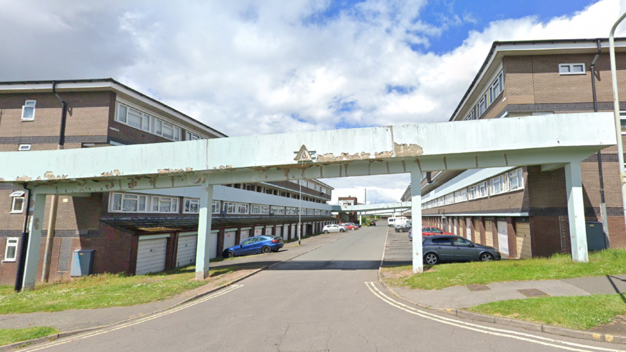 The picture show the current estate which has a tatty concrete entrance to to rows of maisonettes behind with cars parked outside some