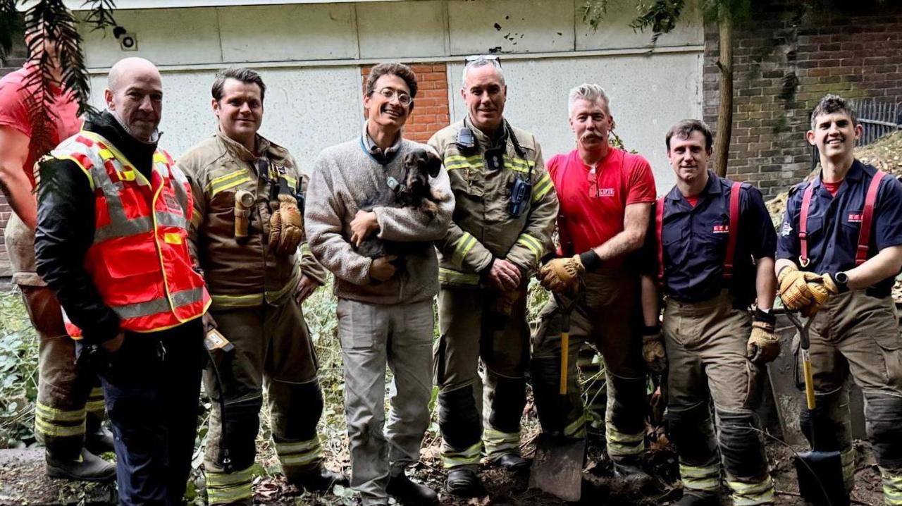 A group of firefighters lined up with the middle one holding the rescued dog. They are standing outside in front of a white wall and some greenery. ~