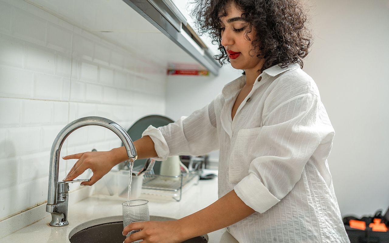 A woman fills a glass with water from a kitchen tap at home, she's wearing a white shirt and the kitchen has white tiles and a dish drying rack just visible behind the sink.