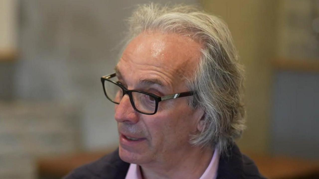 Bartolome Bisbal looks away from the camera while talking. He has a receding hairline and some hair that is grey. He has glasses and wears a navy jumper with a pink shirt underneath.