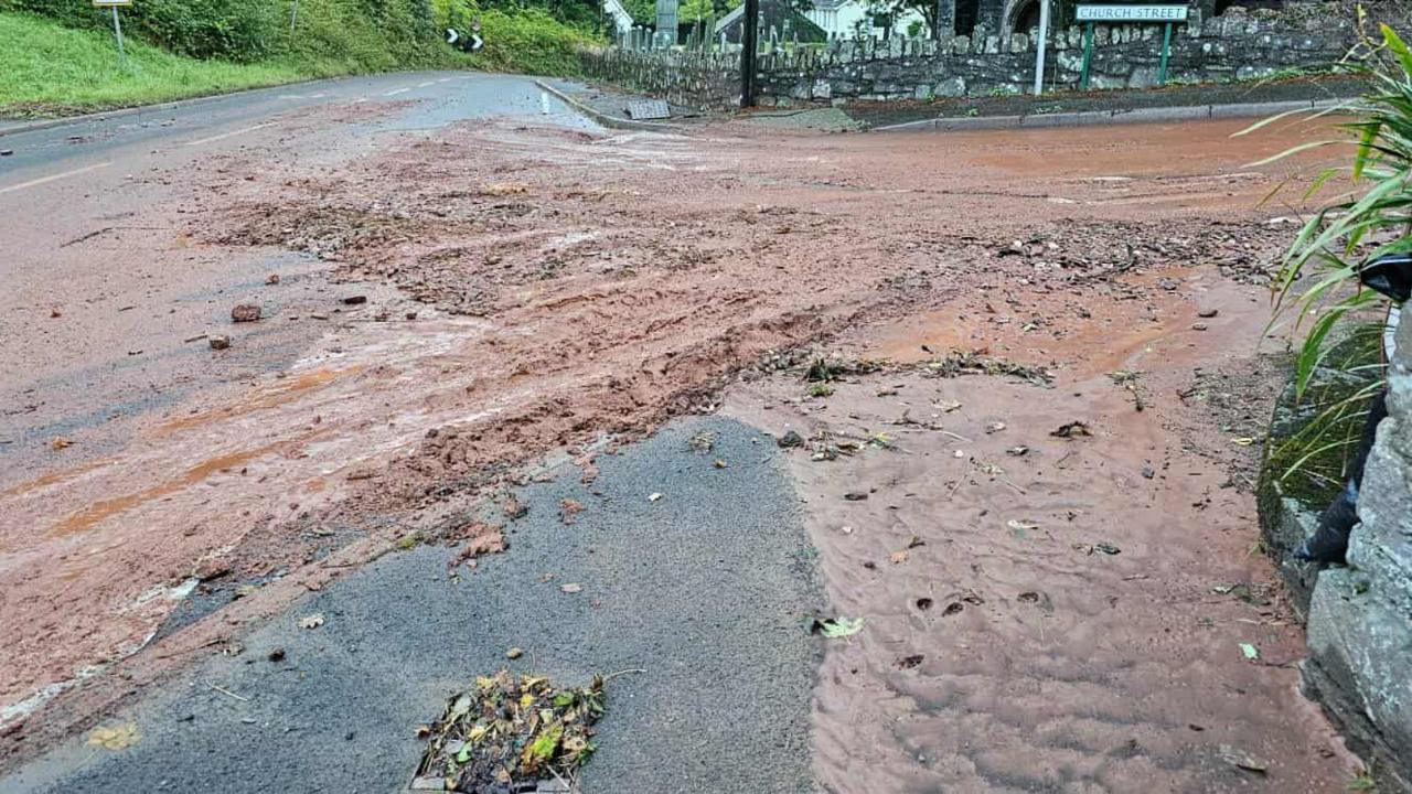 A flooded road mixed with mud besides a pub in Totnes. The water is brown with mud and there is a brick wall on the opposite side of the junction.