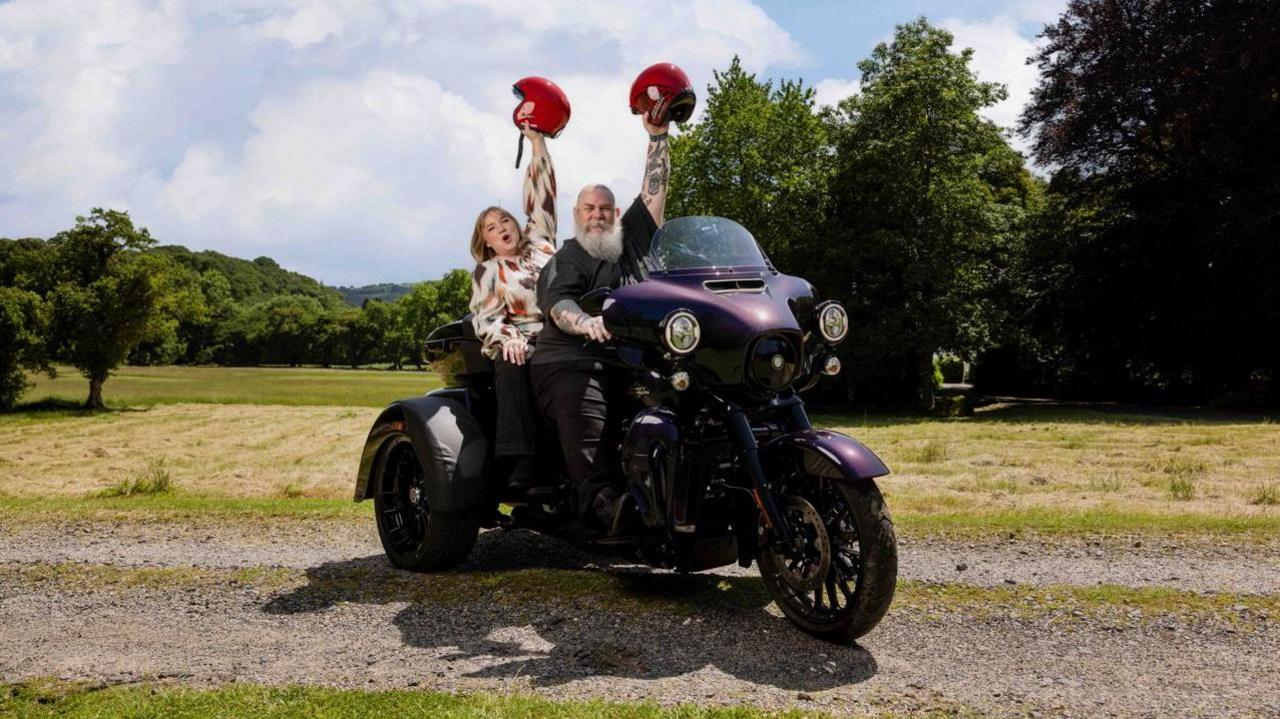 A woman and a man, holding red helmets up, sat on a Harley-Davidson trike bike. Behind them is grass and trees. The sky is blue with white clouds.