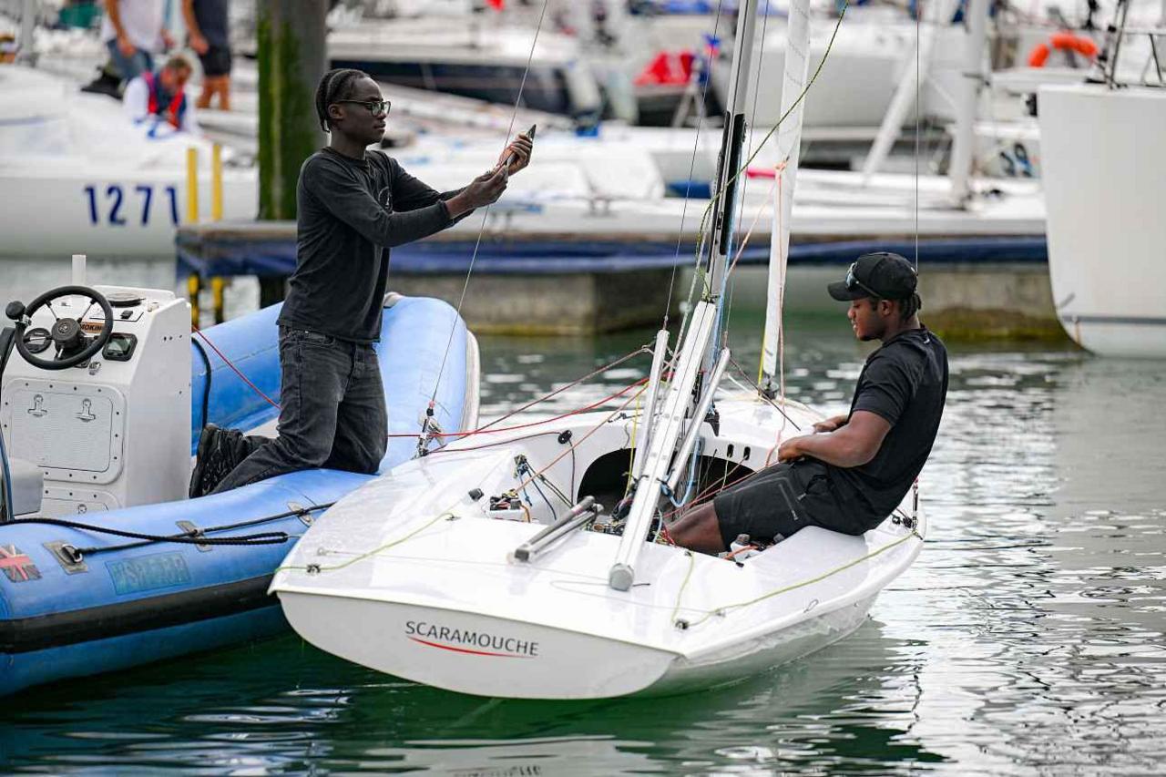 Two sailors prepare Flying 15 SCARAMOUCHE at Cowes Week. A sailor is sitting in the boat while another person kneels on the edge of a blue rib taking his photo with a phone.