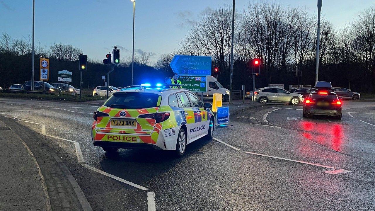 Police car with blue lights flashing stands at entrance to Stanningley Bypass in Leeds, with a queue of traffic in the background.