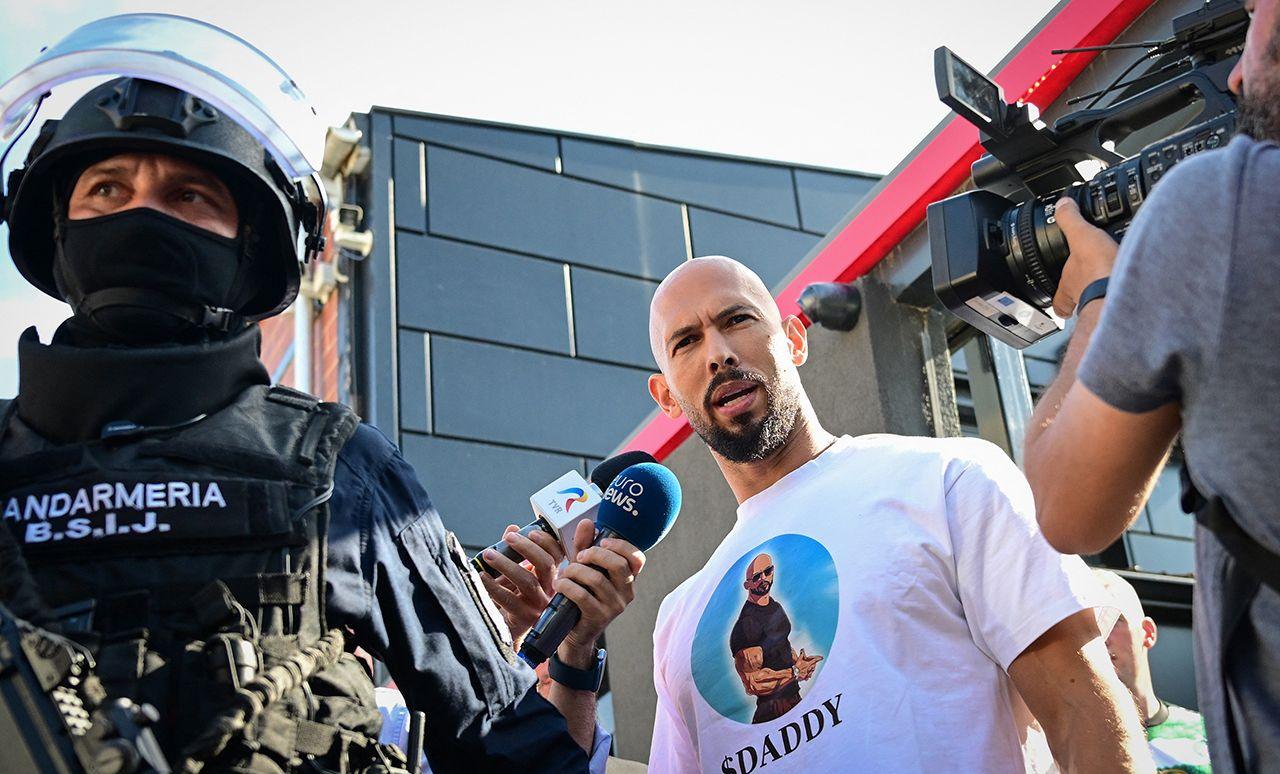 Andrew Tate looks at the camera while being brought in by police, a masked police officer wearing a helmet looks on while reporters gather round Tate, taken outside the Tate brothers' residence in Bucharest on Wednesday night