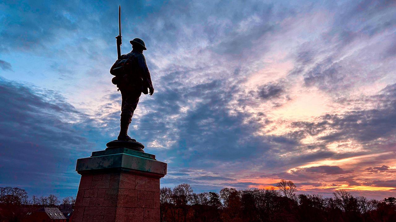 A statue of a soldier carrying a rifle and bayonet on a pedestal in Evesham. It is almost in silhouette as the sun sets in a cloudy sky