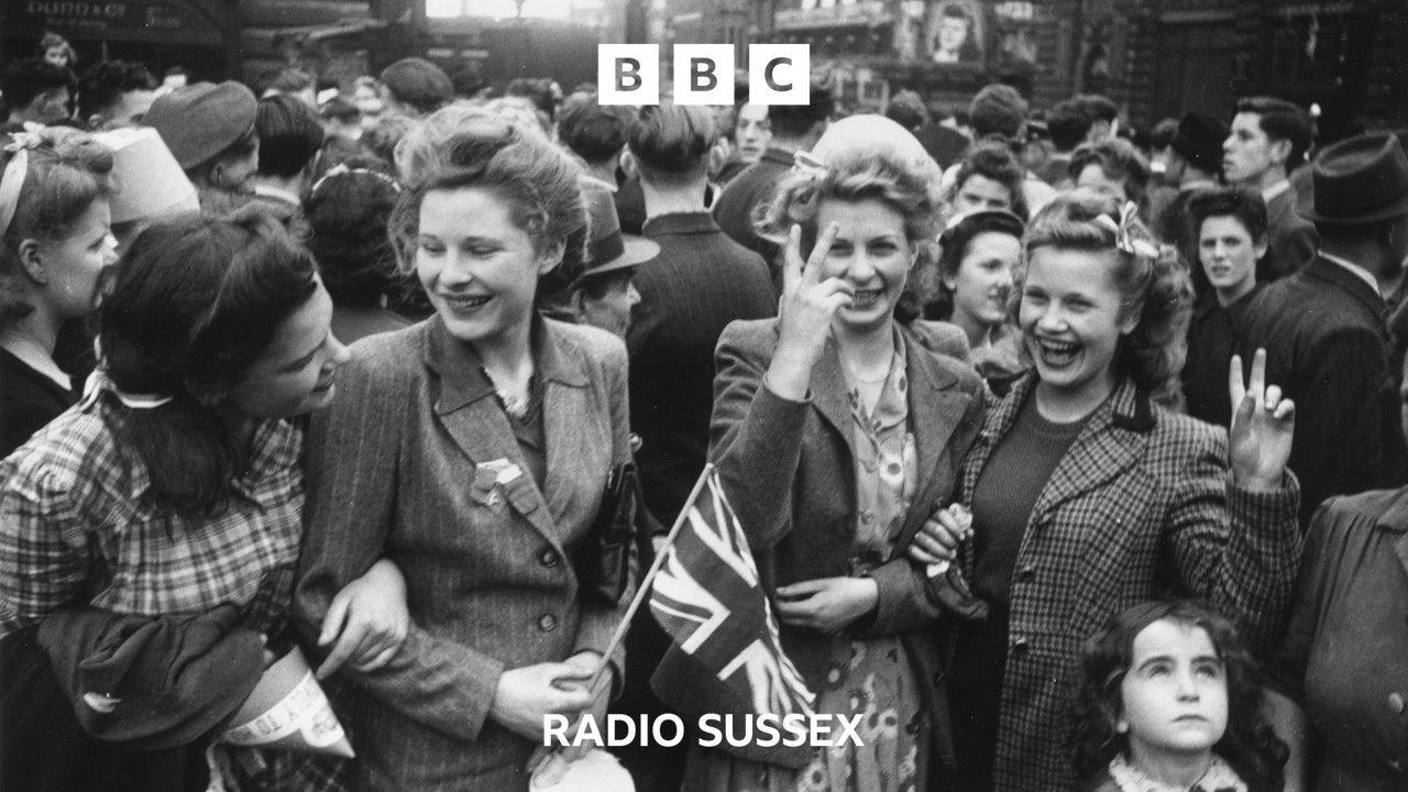 A black and white image of crowds celebrating the end of World War 2. They are outside. In the front are four women, all smiling, one is holding a Union Jack and two have their fingers in a V sign.