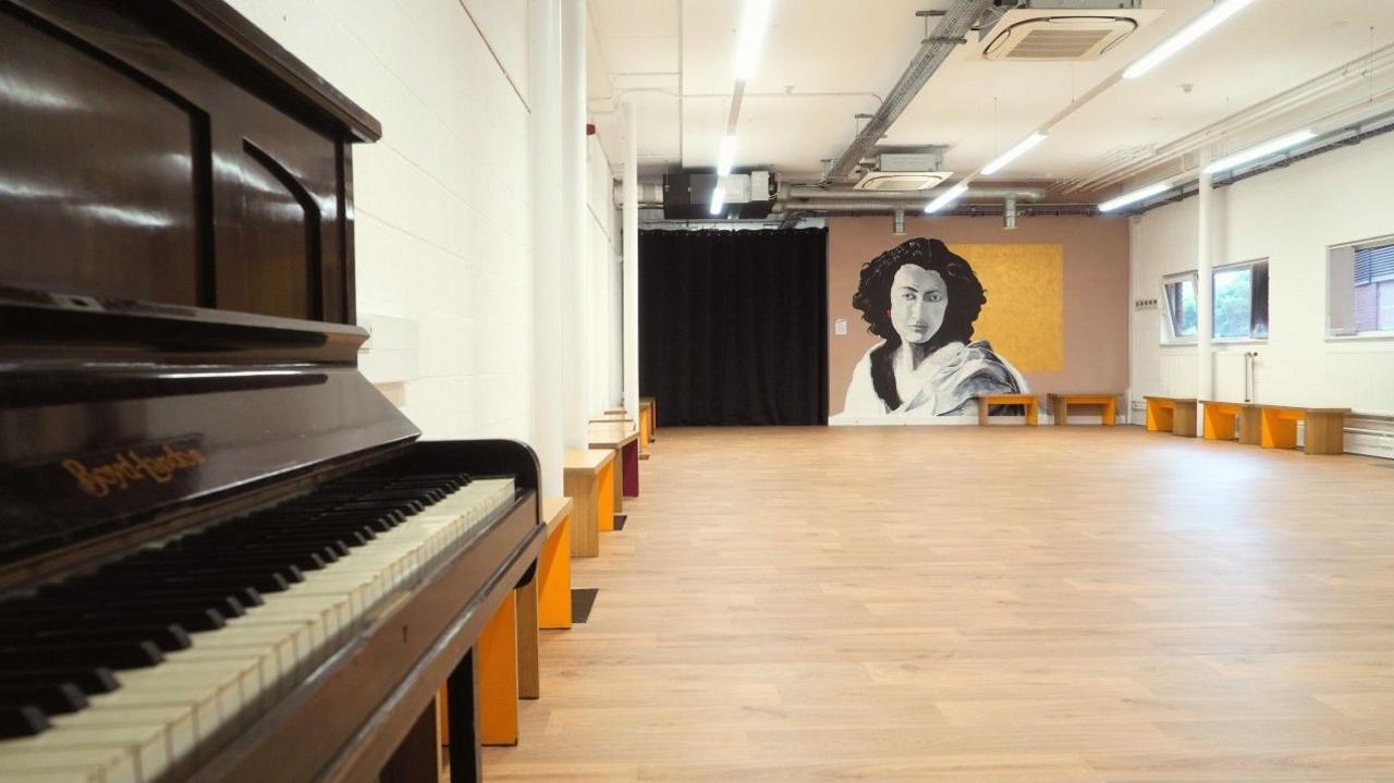 Piano in the foreground in a wood-floored studio with a mural of a woman at the end wall