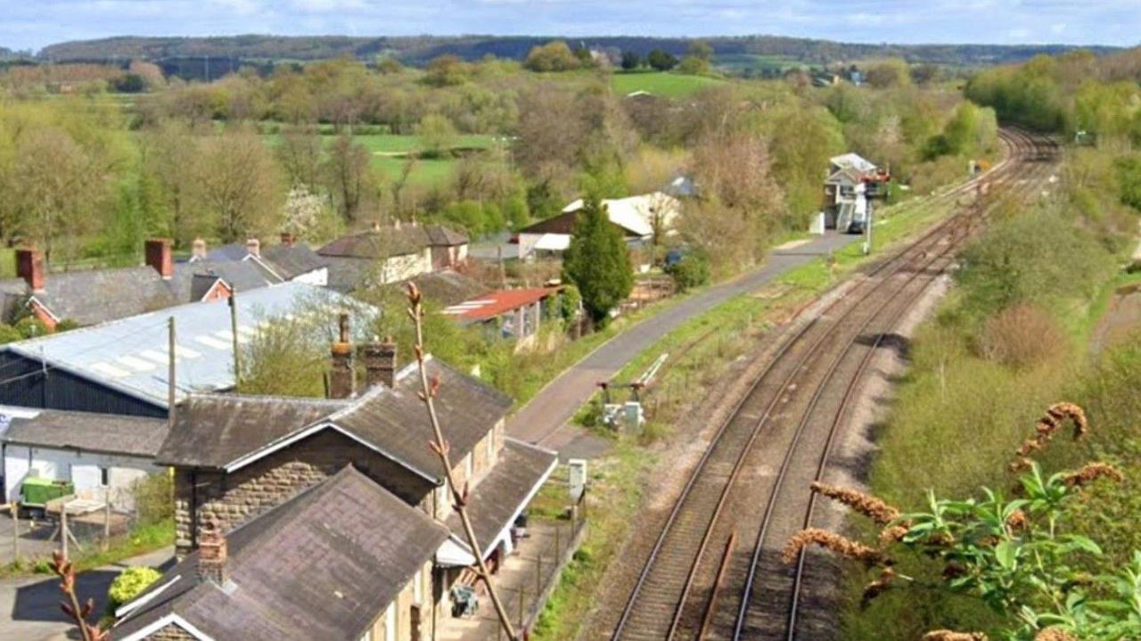 The railway line through Pontrilas, with the image showing tracks on the right of the photo going from the bottom to nearly the top of the image. In the background are fields, trees and a blue sky at the top of the photo.