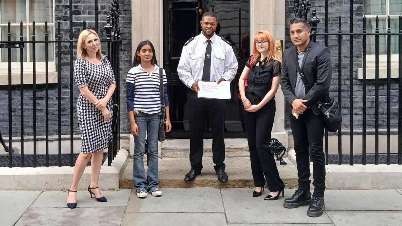 Five people stand in front of the door to 10 Downing Street. A police officer stands in the middle holding a piece of paper. 