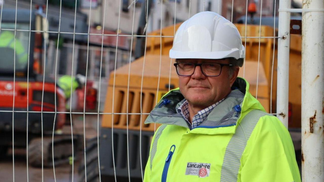 Councillor Rupert Swarbrick stands by a fence covering a building site. He is wear a high-vis council jacket and a white hard hat. 