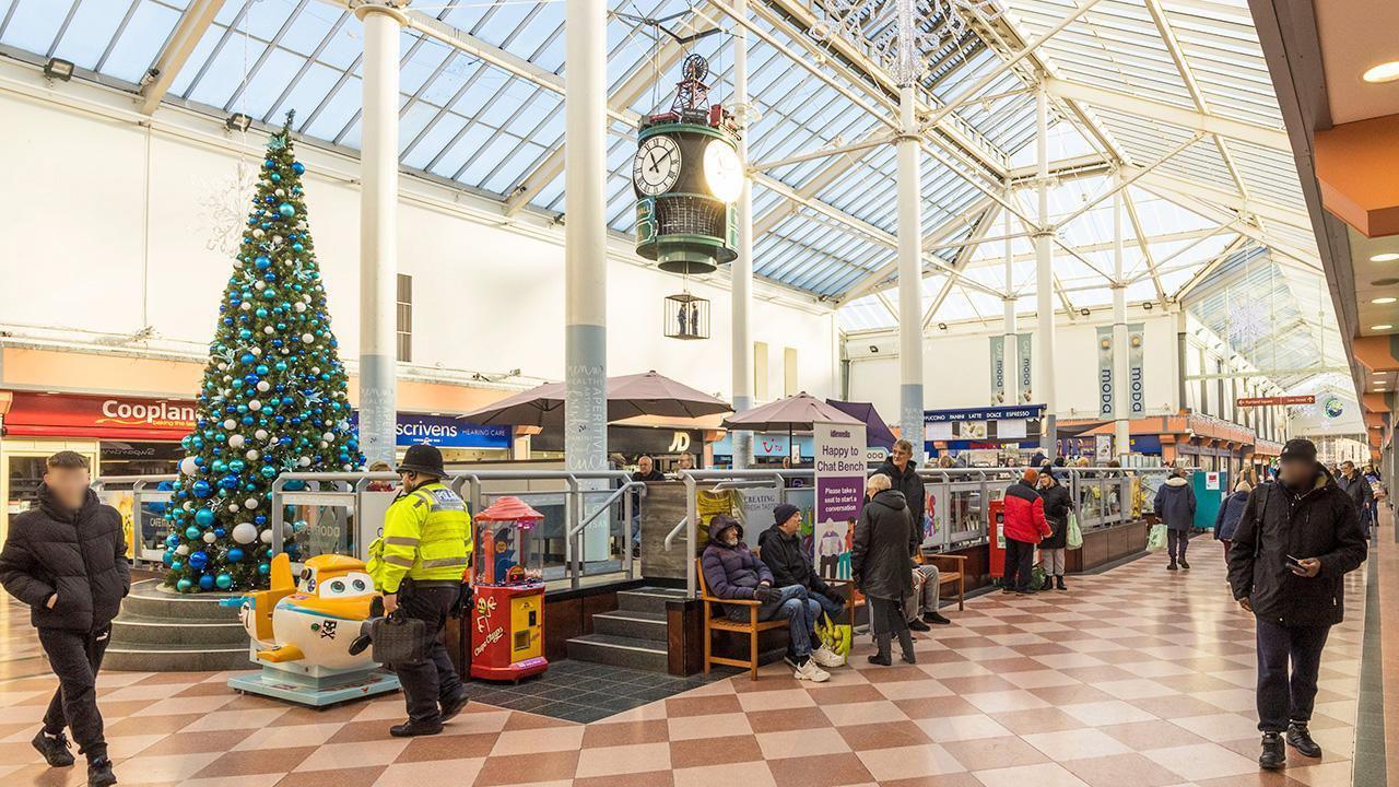 A group of shoppers walking through Idlewells shopping centre. There is a Christmas tree with silver and blue baubles in the background. 