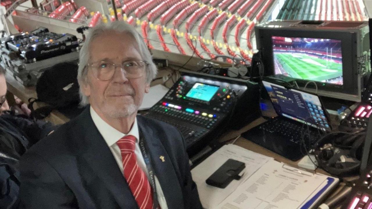 Iestyn Garlick yn gwenu at y camera gyda a seddi gwag y stadiwm a sgrin y tu ol iddo. Mae e'n gwisgo sbectol, tei coch a siwt.