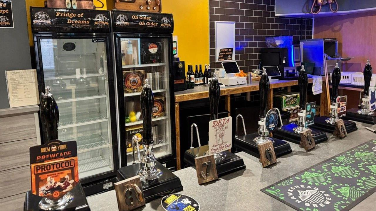 A bar, with beer mats placed along the surface and fridges and counters behind. The walls are a mixture of dark yellow and grey tiles in a brick effect. There are seven handles to pull pints and tills in the background too.
