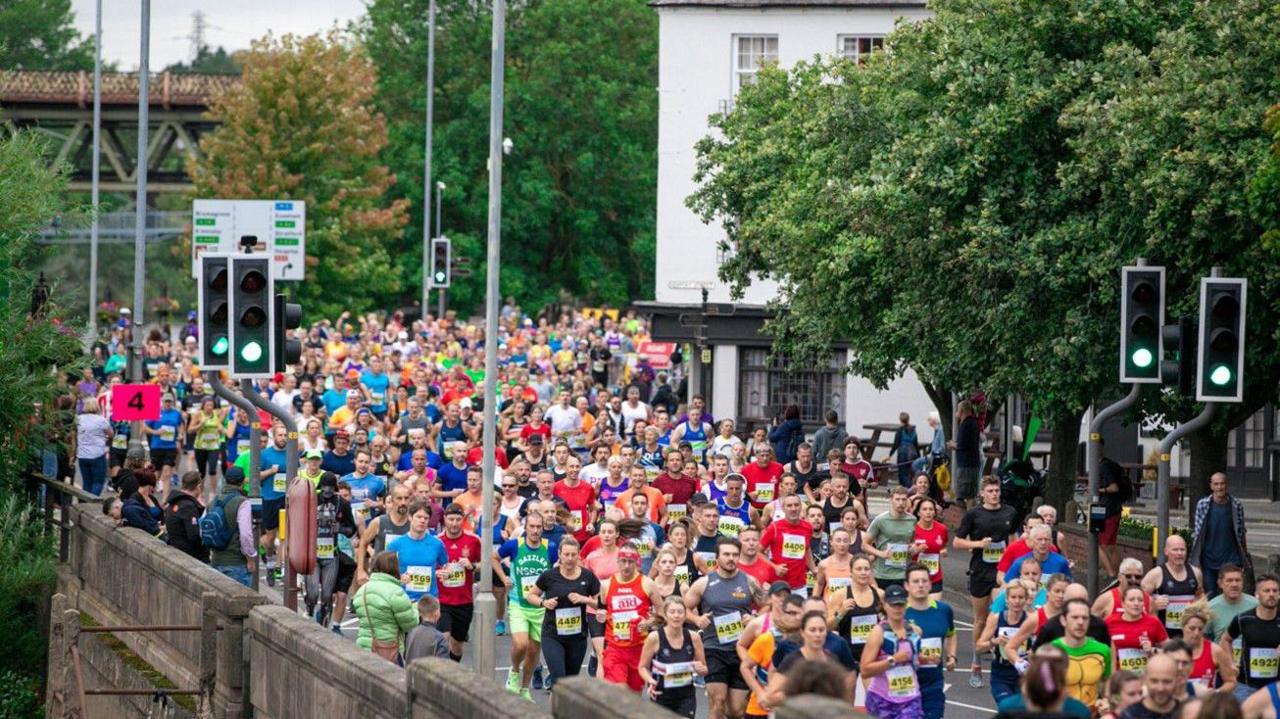 An image from 2022 shows more than 100 runners in the bottom half of the photo. Trees, a white building and lampposts are above them on the photo.