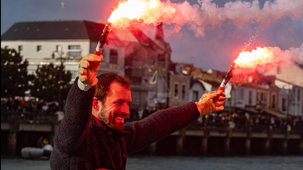 A man holding red flares in each hand smiles with a crowd in the background.