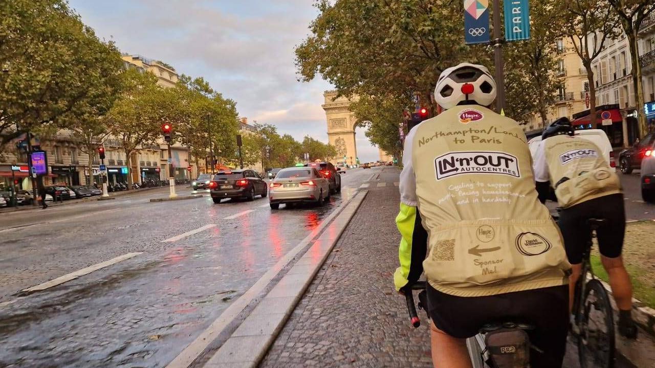 Two men in green cycling vests and cycling shorts approach the Arc de Triomphe in Paris on bikes. The tree-lined road is wide with cars on either side and buildings behind and a large stone arch in the distance.