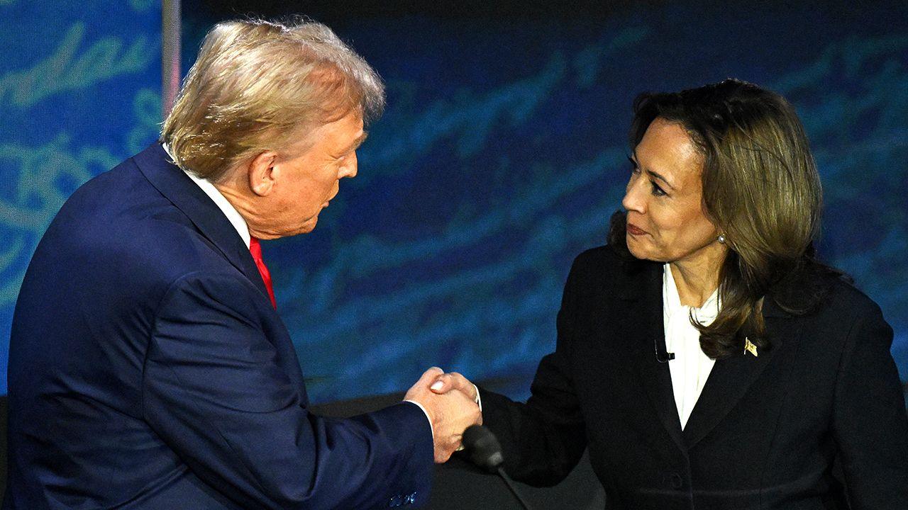 Donald Trump and Kamala Harris shake hands while smiling courteously at each other, both wearing suits, against a blue backdrop on stage in Philadelphia in September