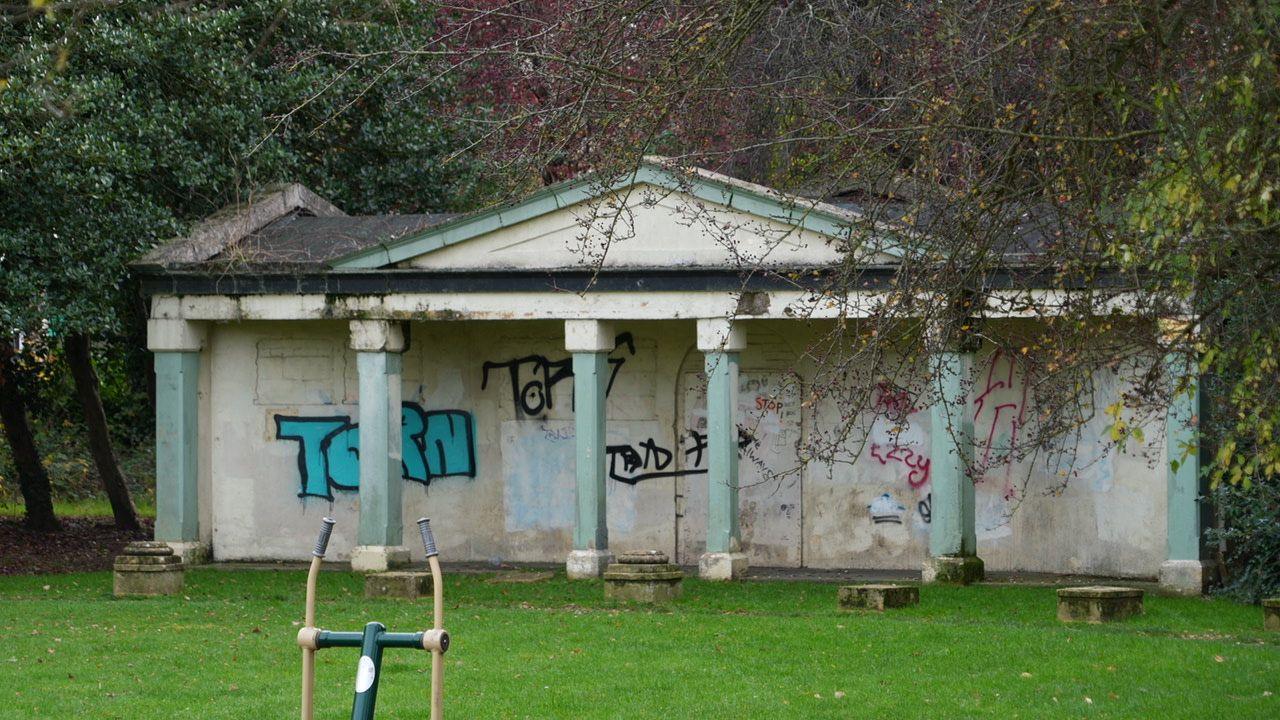 A pale stone pavilion fronted by six mint green columns. The pavilion is boarded-up, with the boards covered in graffiti. The area in front of the pavilion is laid to lawn.  