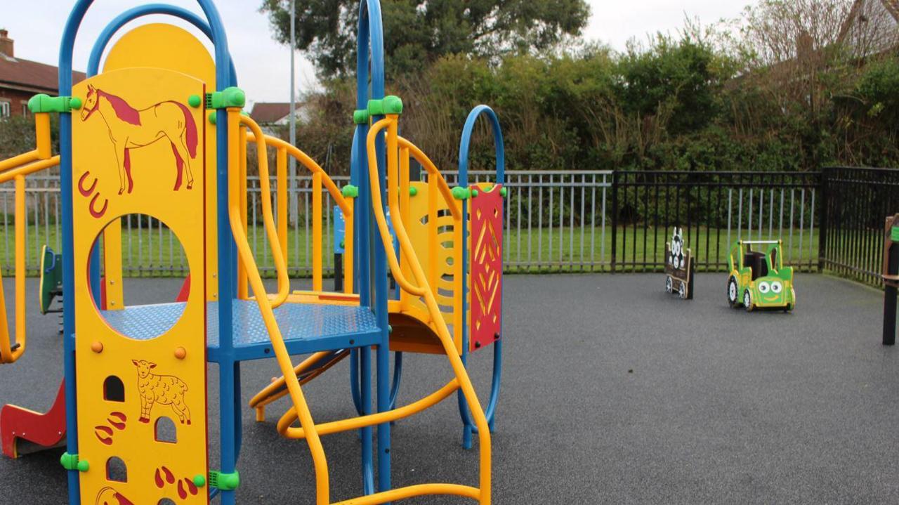 An enclosed play area with smooth tarmac flooring. A yellow, blue, green and red climbing frame can be seen on the left. It has a drawing of a horse and a sheep on it. On the right is a green cartoon car with eyes and a smiling mouth.