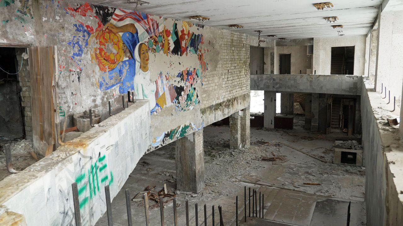 Interior view of the Palace of Culture, showing bare brick walls and concrete floors. Part of a colourful mural remains, and loose tiles and other debris are on the ground.