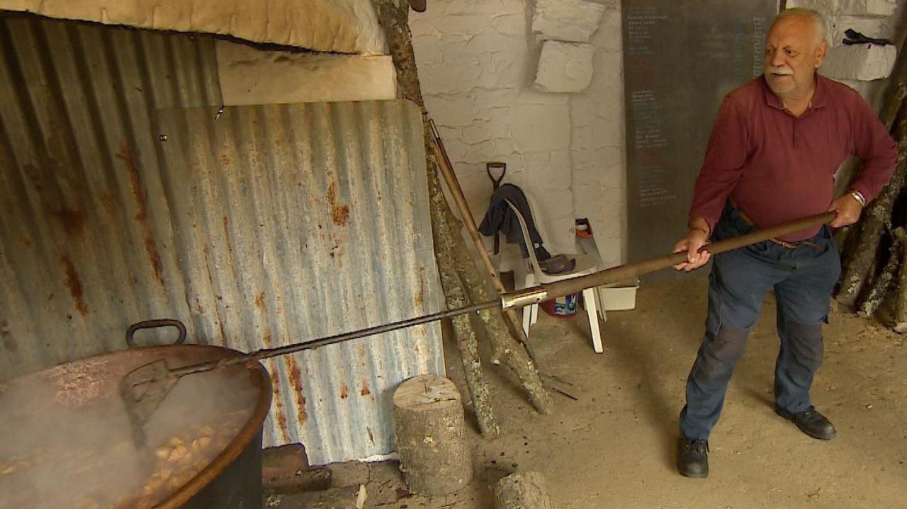 Jean Priaulx wearing a burgundy polo shirt and navy blue trousers uses a long stick to stir the contents of a large pan above a log fire.