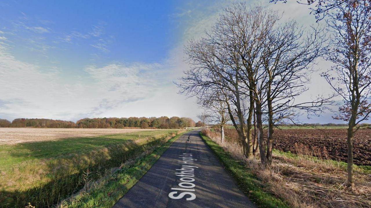 The image shows a country road with fields on the left and trees on the right. The trees have no leaves. The sky is blue with light clouds. 