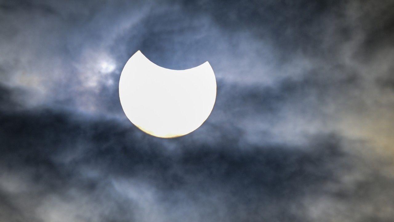 The Moon moves over the edge of the Sun in a sky filled with blue, white, black and brown coloured clouds 