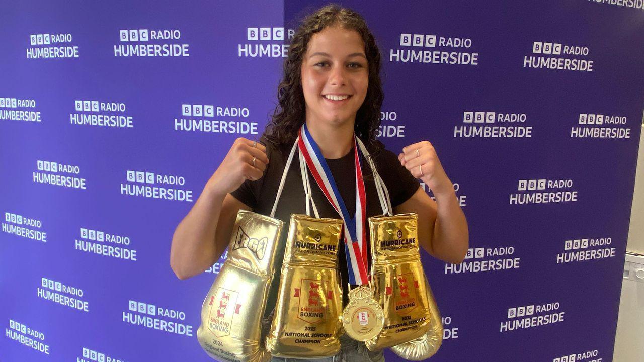 A young girl with dark brown curly hair is smiling at the camera and holding up both her fists. She is wearing a black top and around her neck are three gold boxing gloves and a gold medal with blue, white and red stripes on the ribbon. In the background is a purple wall which repeatedly says 'BBC Radio Humberside'.