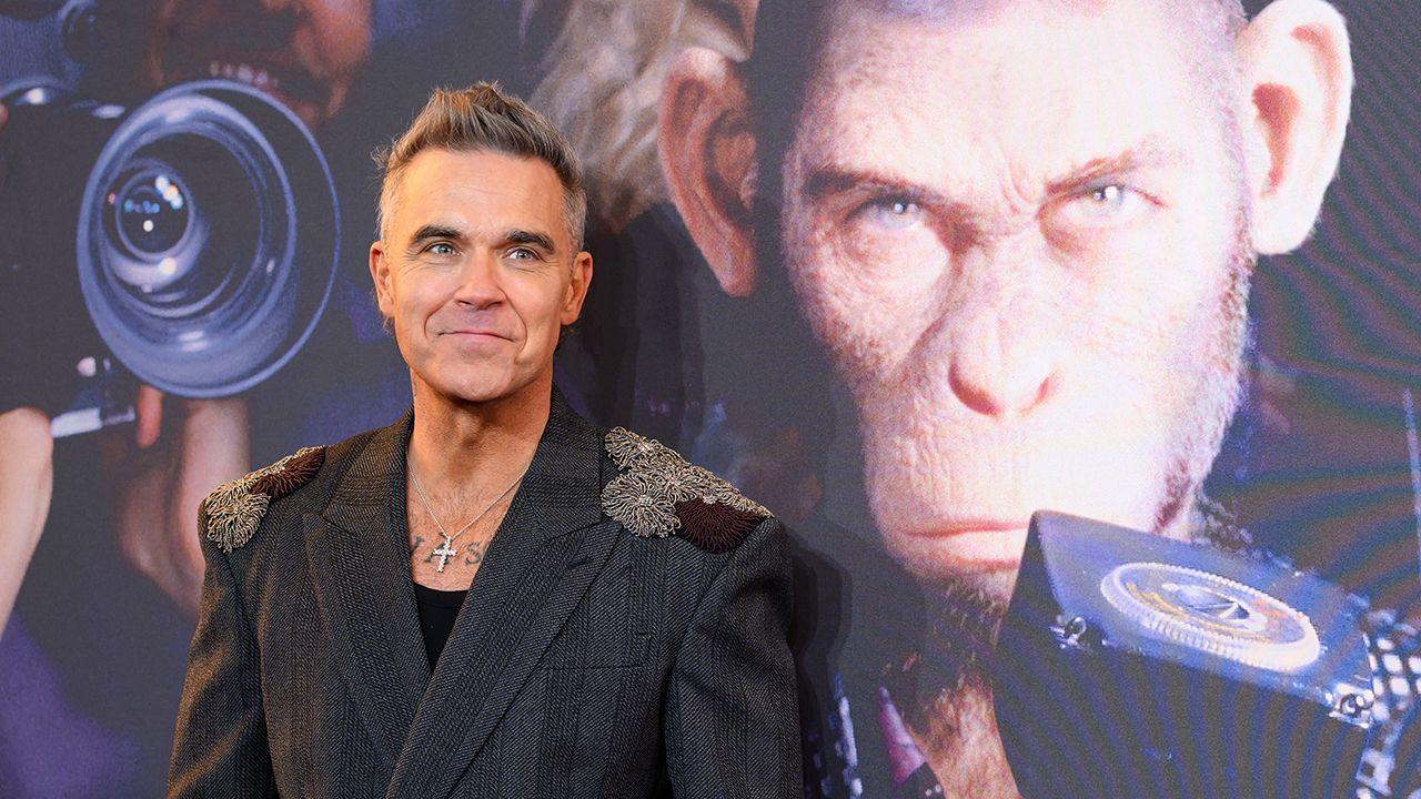 Robbie Williams stands smiling wearing a black suit in front of the promo image of a chimpanzee playing him in a new biopic named Better Man, at the European premier in Leicester Square on 27 November.