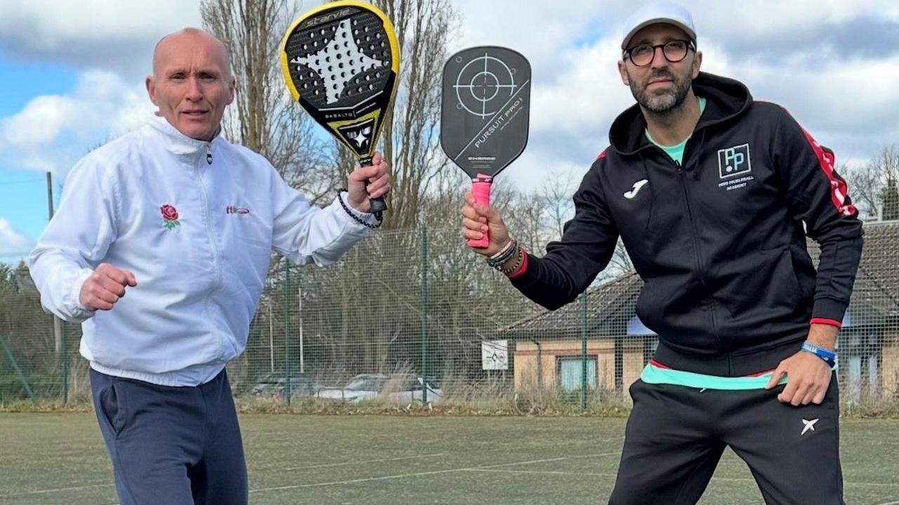 Alan wearing a white track suit jacket and grey trousers and Pep wearing a black track suit- both holding padel tennis rackets 