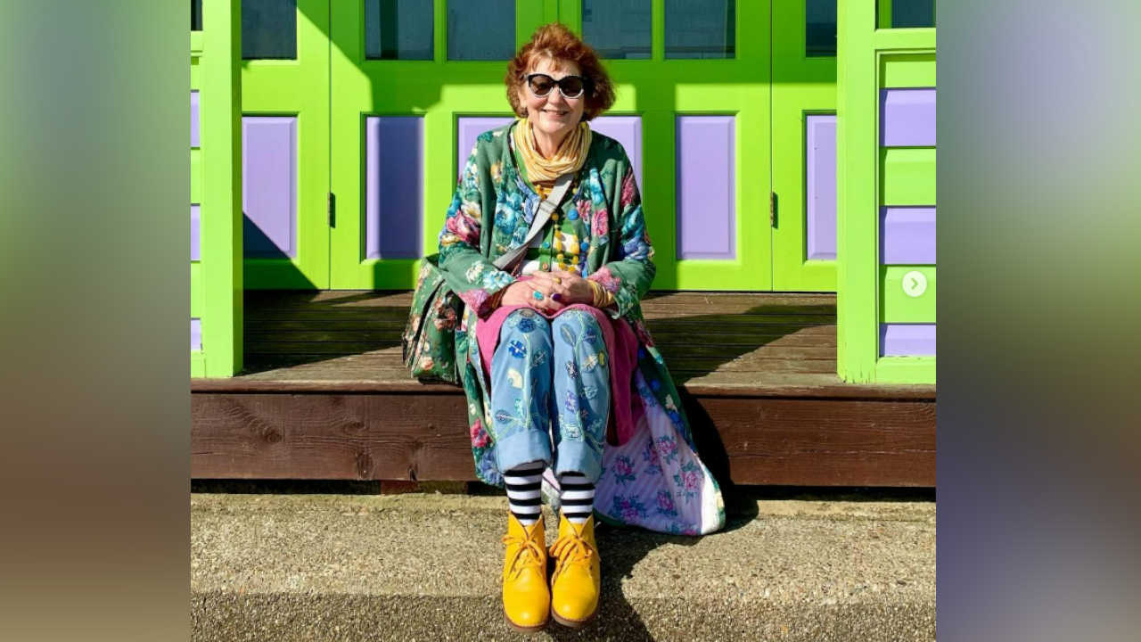 Marcia Riddington with short red hair and black sunglasses sits on a ledge outside a bright purple and green beach hut. She is wearing a green dress with flowers sewn into it and blue trousers with yellow boots.