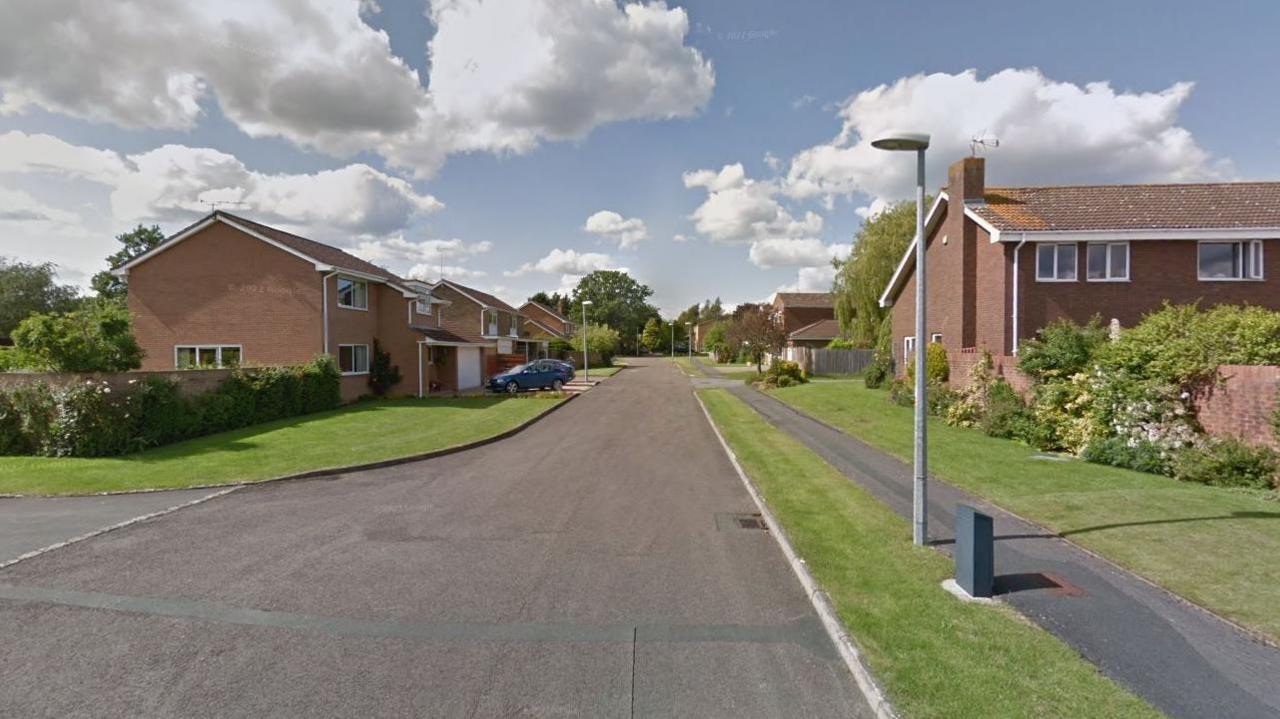 A google maps street view of Oakbourne Park. It is a quiet residential street with brick houses and a wide road. There is a pavement on the right with grass either side. 