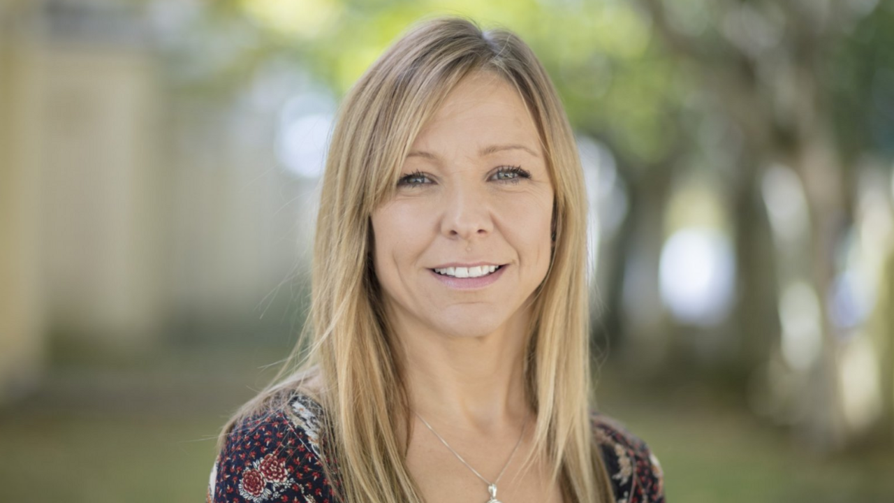 Claire Chick, who is smiling and has straight, shoulder-length hair. She is in an outdoor setting and is wearing a patterned top with a subtle necklace.