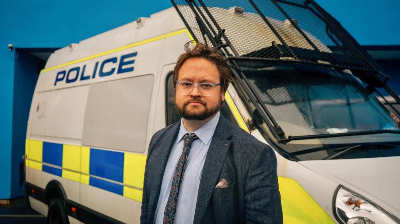 Matt Storey standing in front of a police van. He has brown hair and a beard and is wearing glasses, a navy blazer and a light blue shirt with a dark tie.