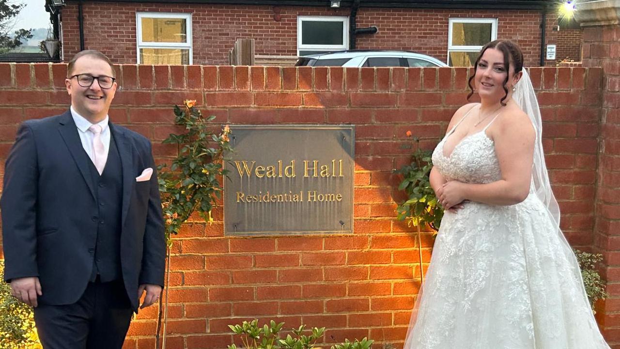 Jack and Lauren are pictured outside the care home in front of its entrance sign in their wedding attire. The care home entrance sign sits on a red brick wall and is written in gold writing against a square black backdrop.