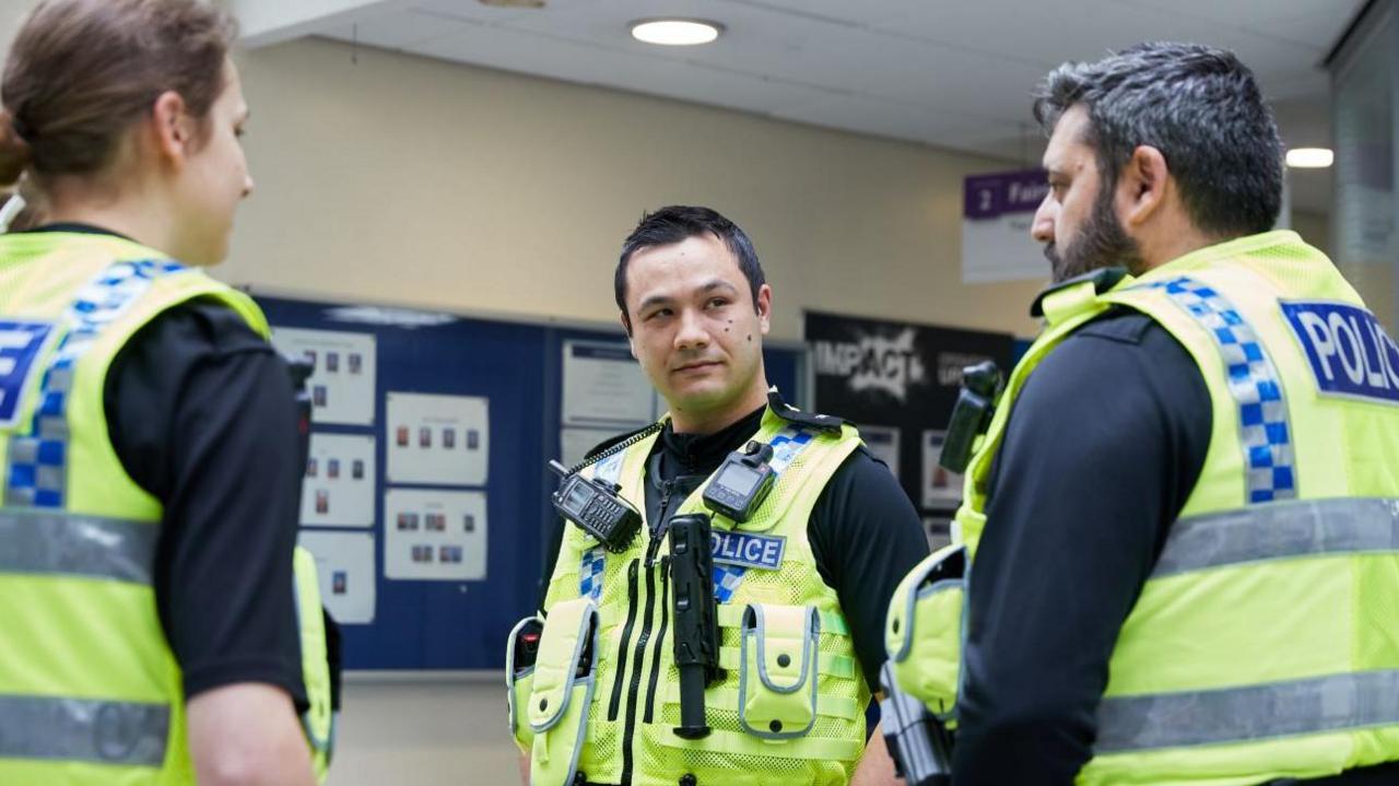 Three police officers, a woman and two men, stand looking at each other in a hall inside a police station. Each is wearing a green high-vis police jacket. 