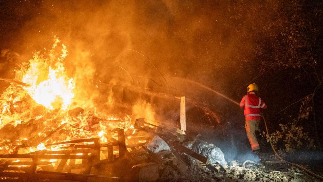 A firefighter tackles a bonfire which has got out of control and spread to nearby trees. He points a water hose at the flames.