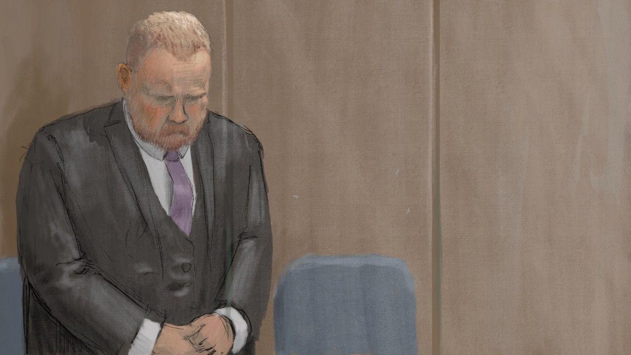 Robert Bush stands in the dock at Hull Crown Court wearing a formal dark suit, white shirt and mauve tie. He has his head down and his hands clasped in front of him. 