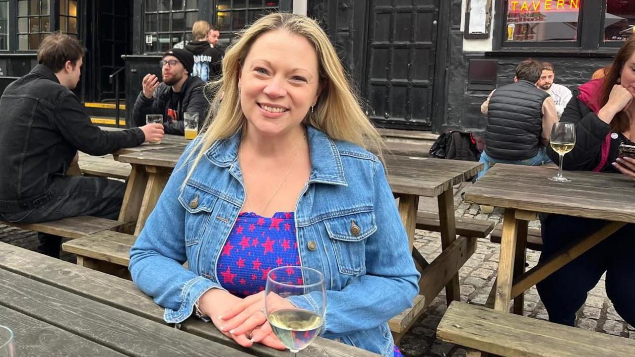 Toni sitting at a bench in Bristol city centre smiling with a glass of white wine