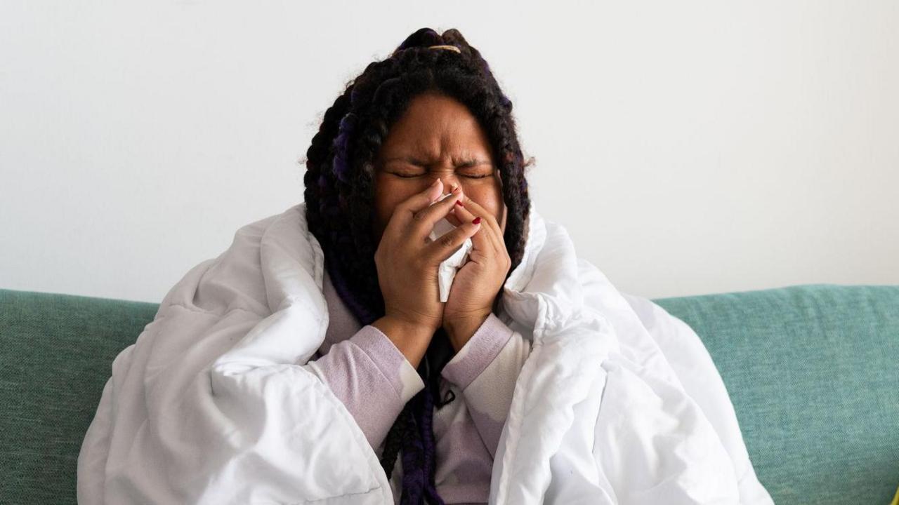 Young woman sitting on the sofa at home, covered with a blanket, blowing her nose, suffering from a cold or flu.